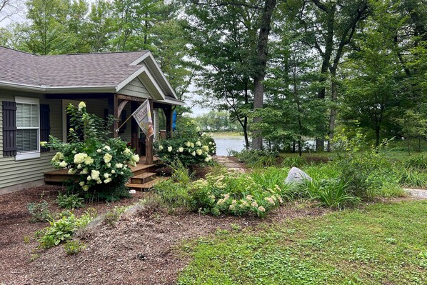A house with a porch, surrounded by a garden with hydrangeas and a view of a lake. - Fish, Hike & Hunt: Lakeside Newaygo Cottage (Newaygo)