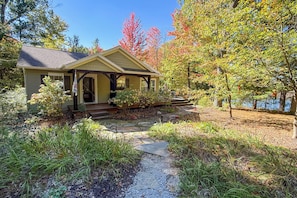 A two-story house with a porch, a stone pathway leading to the entrance, and a garden with various plants and shrubs. - Fish, Hike & Hunt: Lakeside Newaygo Cottage (Newaygo)