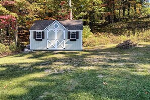 A small, single-story shed with a gabled roof and a central door, surrounded by a grassy area and trees with autumn foliage. - Fish, Hike & Hunt: Lakeside Newaygo Cottage (Newaygo)
