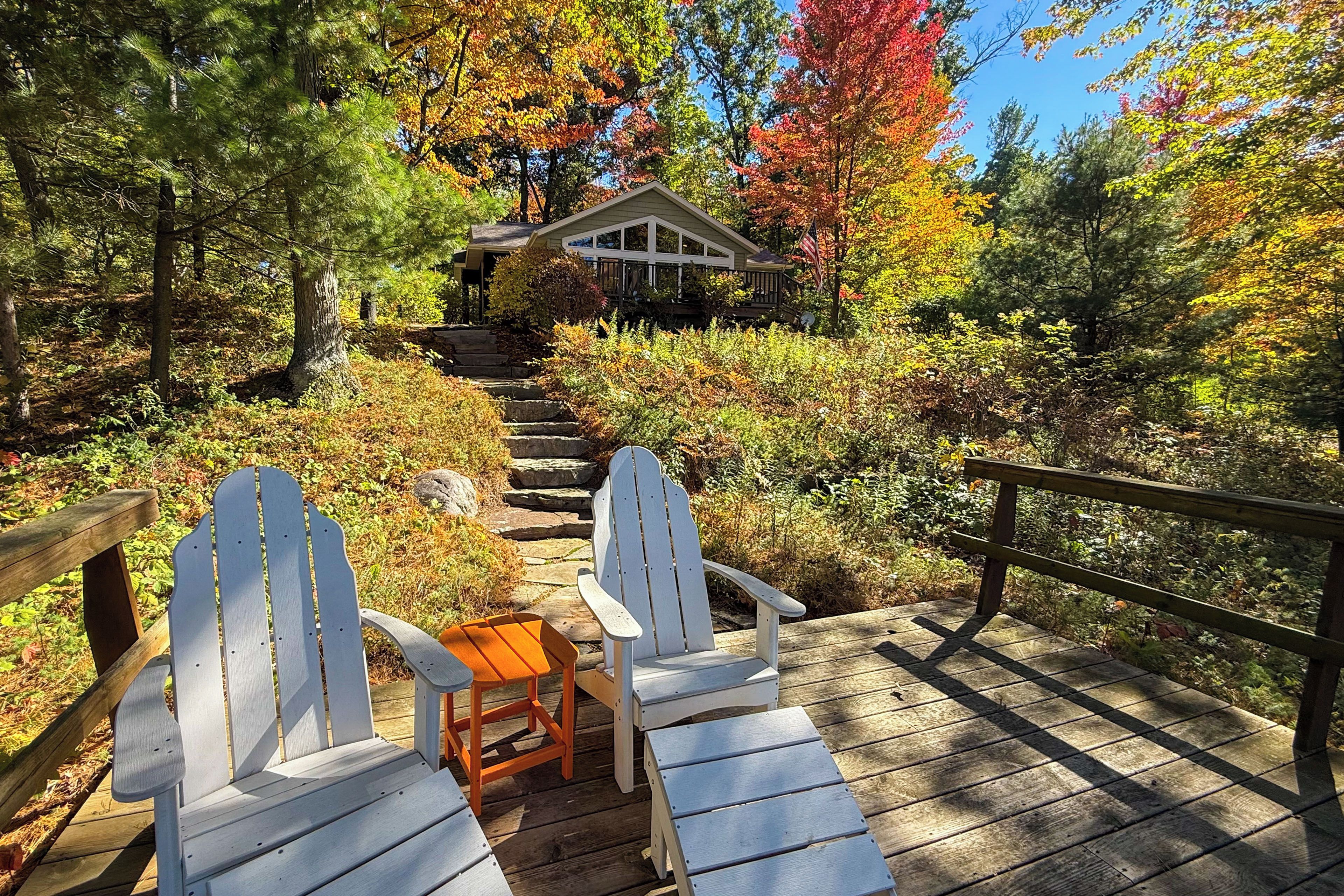 Une terrasse en bois, agrémentée de chaises Adirondack et d’une petite table, qui mène à une maison dotée d’un porche.
