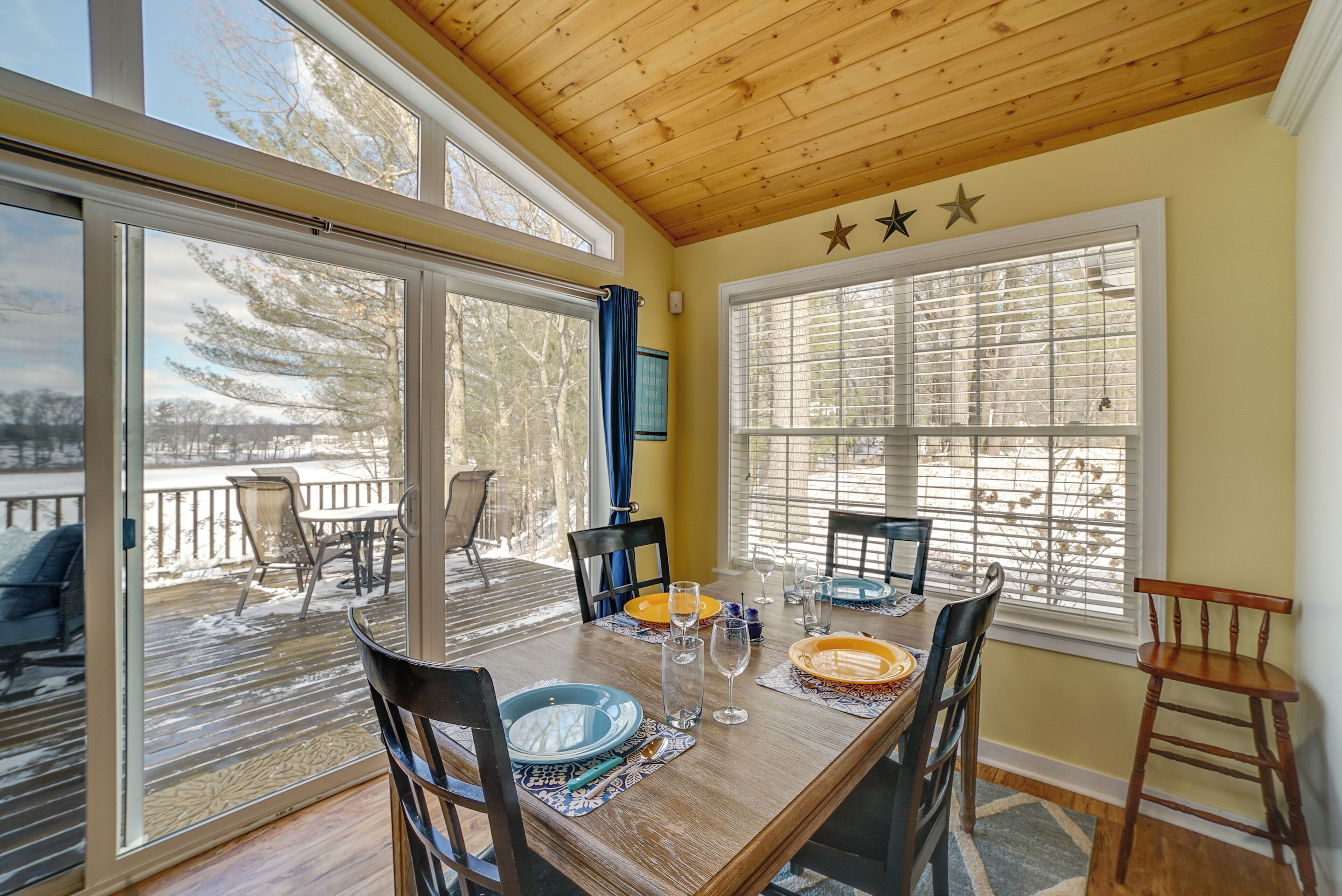 A dining area with a wooden table set for four, surrounded by chairs, with large windows offering a view of a snowy deck and trees.