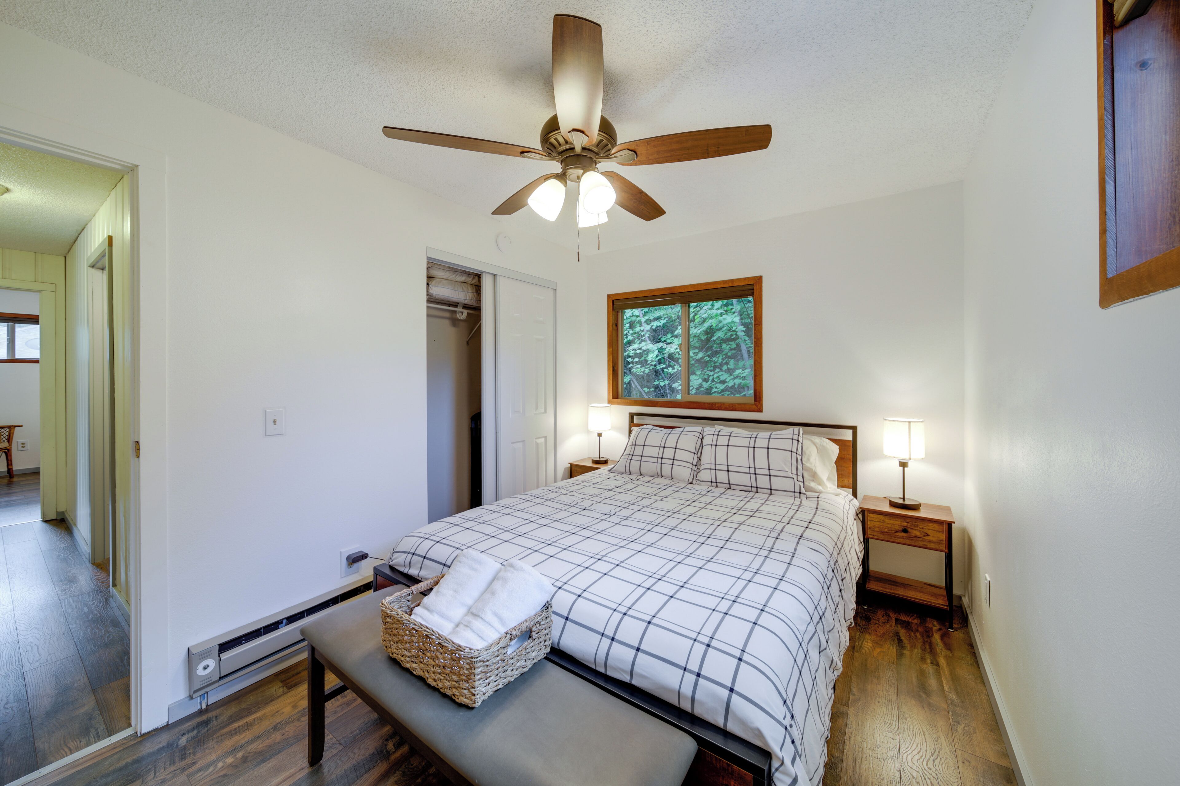 A bedroom with a bed, a ceiling fan, a window, a bedside table with a lamp, and a bedside basket.