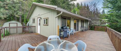 A wooden deck with a glass table and chairs, a barbecue grill, and a small building with a door and windows.