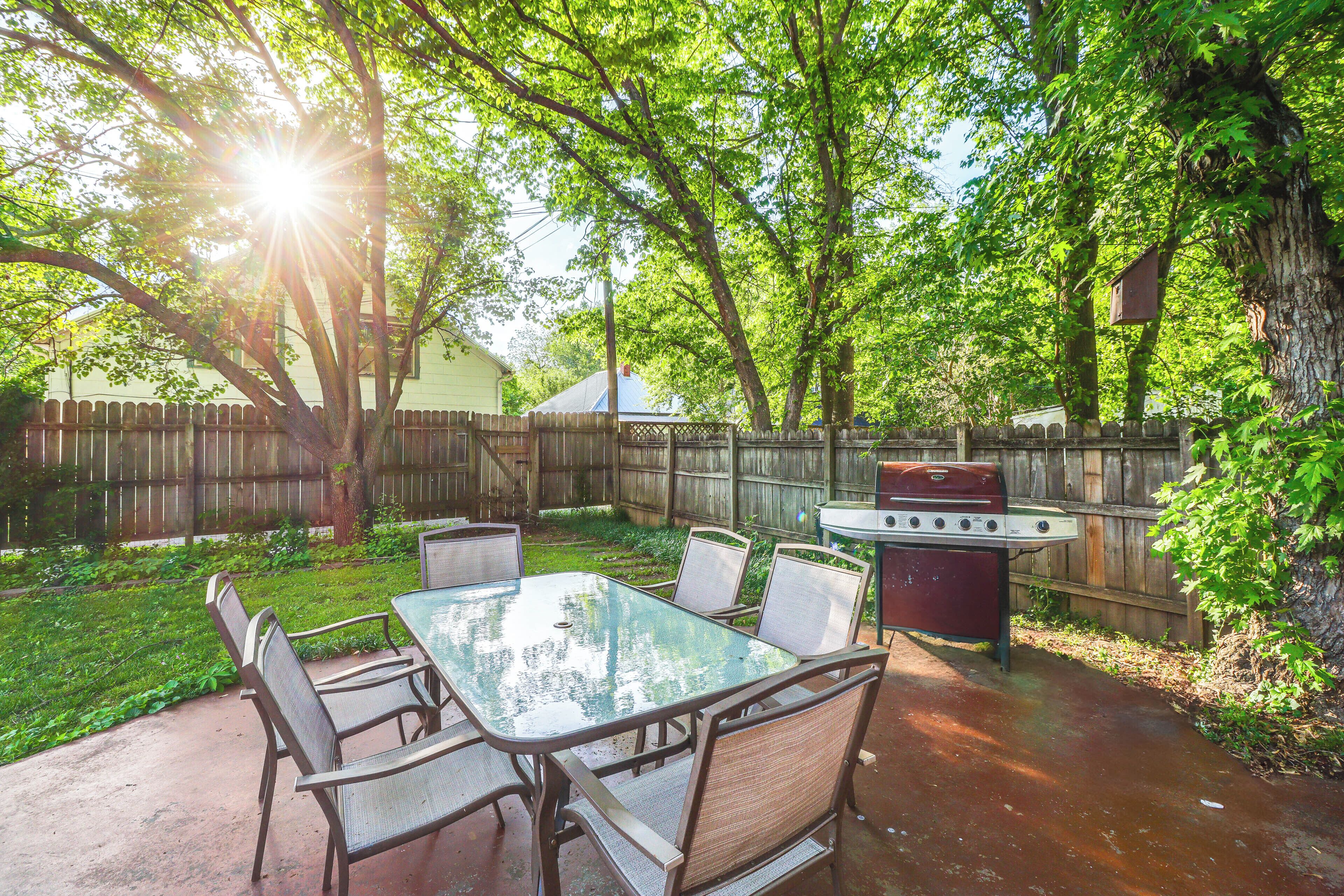 An outdoor patio with a glass-top table and metal chairs, surrounded by trees and a wooden fence.