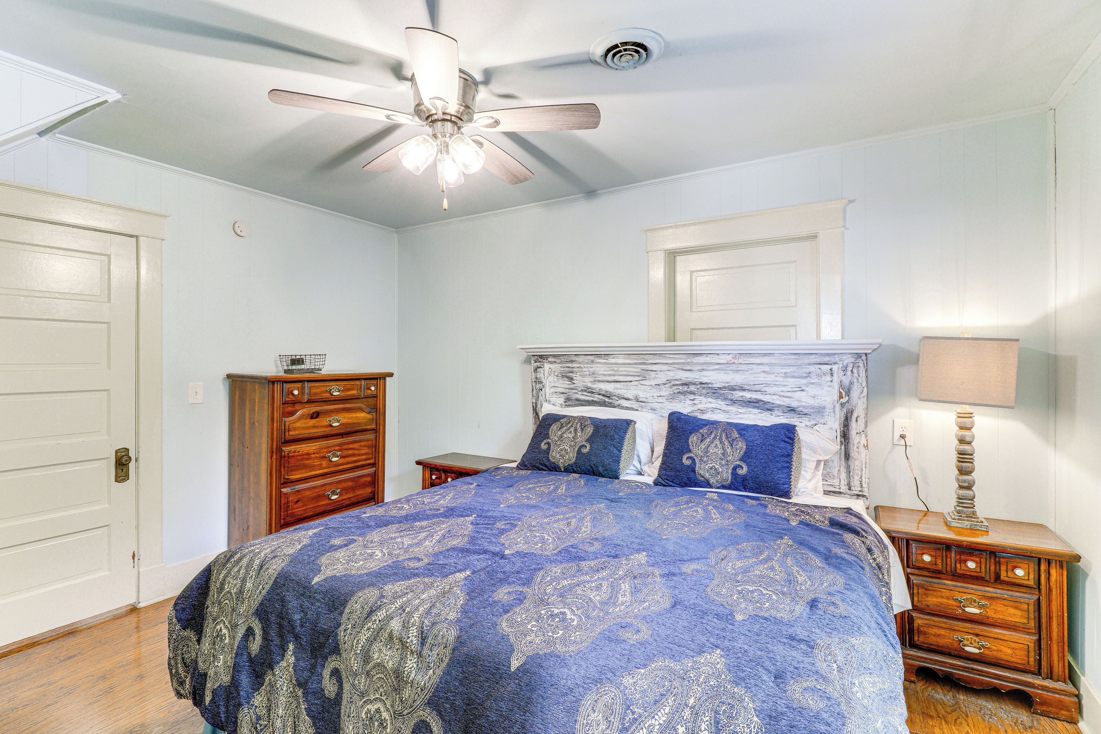 A bedroom with a bed, a wooden dresser, a nightstand, a ceiling fan, and a wall-mounted lamp.