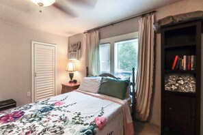Un dormitorio con cama, mesita de noche, lámpara, ventana con cortinas y estantería.