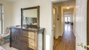 Une chambre avec une commode, un miroir et une chaise. Un couloir mène à une autre pièce.