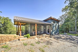 A modern house with a covered patio, a dirt driveway, and a forested area.