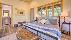 A bedroom with a large bed, a wooden bench, a nightstand, and a window with a view of trees. - Contemporary Home w/ Deck & Mountain Views! (Waynesville)
