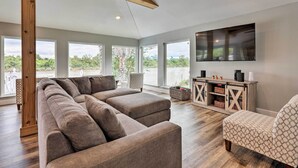 A modern living room with a sectional sofa, a patterned armchair, a wooden ceiling beam, a large TV, and a wooden cabinet with baskets.