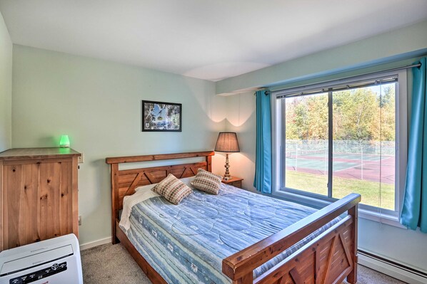 A bedroom with a wooden bed, a nightstand with a lamp, a window with curtains, and a view of a tennis court outside. - Comfortable Dover Apartment: Walk to Mt Snow! (West Dover)