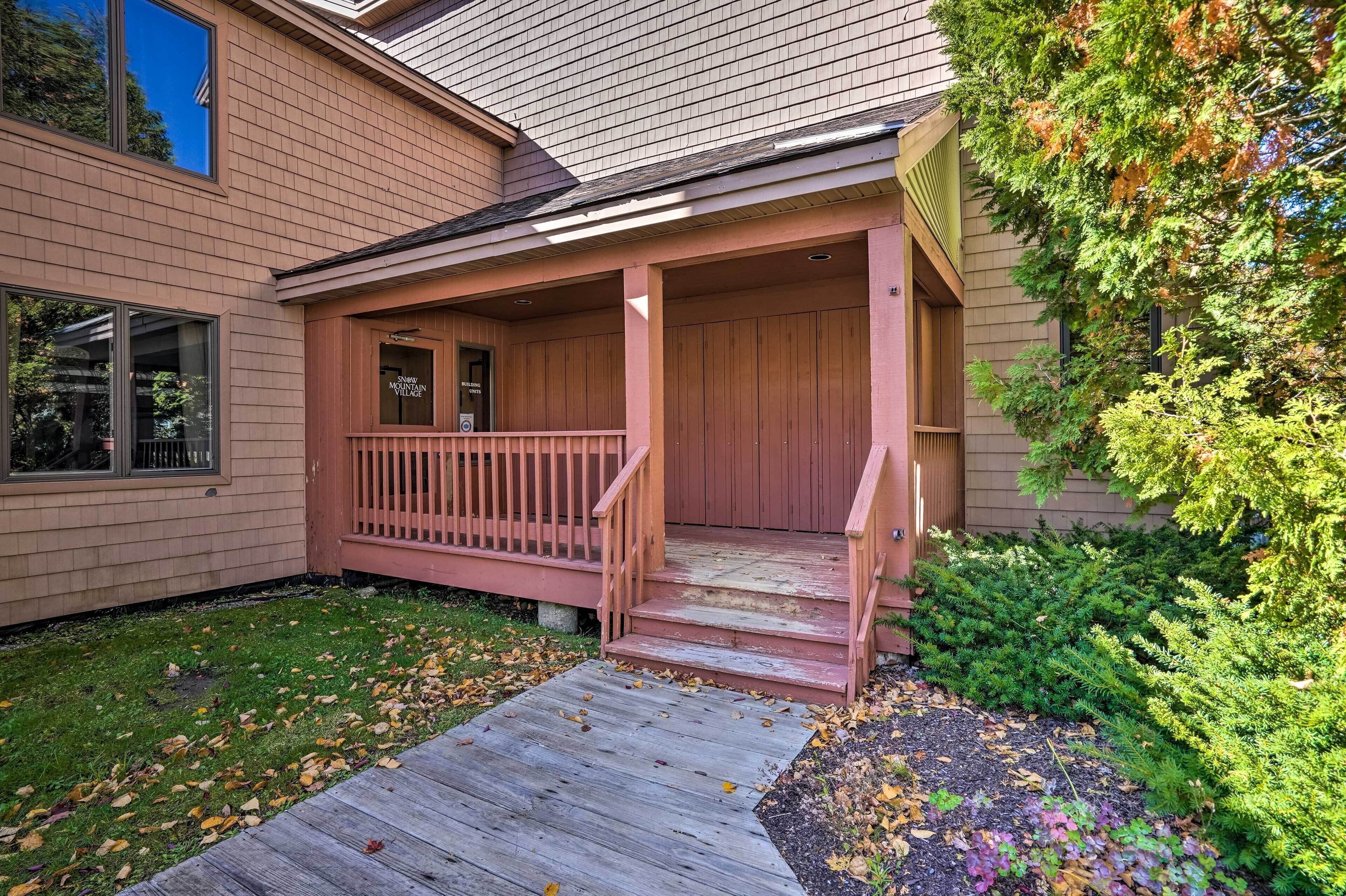 A house with a wooden porch, a front door, and a small garden area with grass and shrubs.