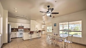 A modern kitchen with white cabinets, a central island with stools, a ceiling fan, and large windows with a view of greenery. - Inviting Bakersfield Home w/ Spacious Yard! (Bakersfield)