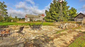 A rustic outdoor setting with a stone patio, metal chairs, a metal table, a wooden barn, and a windmill. - Leelanau Country Cottage ~ 4 Mi to Suttons Bay! (Suttons Bay)