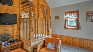 A wooden cabin interior with a television, a desk with a plant, a chair with a cushion, a window with curtains, and a wooden staircase. - Leelanau Country Cottage ~ 4 Mi to Suttons Bay! (Suttons Bay)