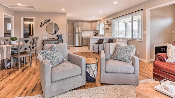 A modern living room with grey sofas, a coffee table, and a red leather sofa. A kitchen with white cabinets and stainless steel appliances is visible in the background.