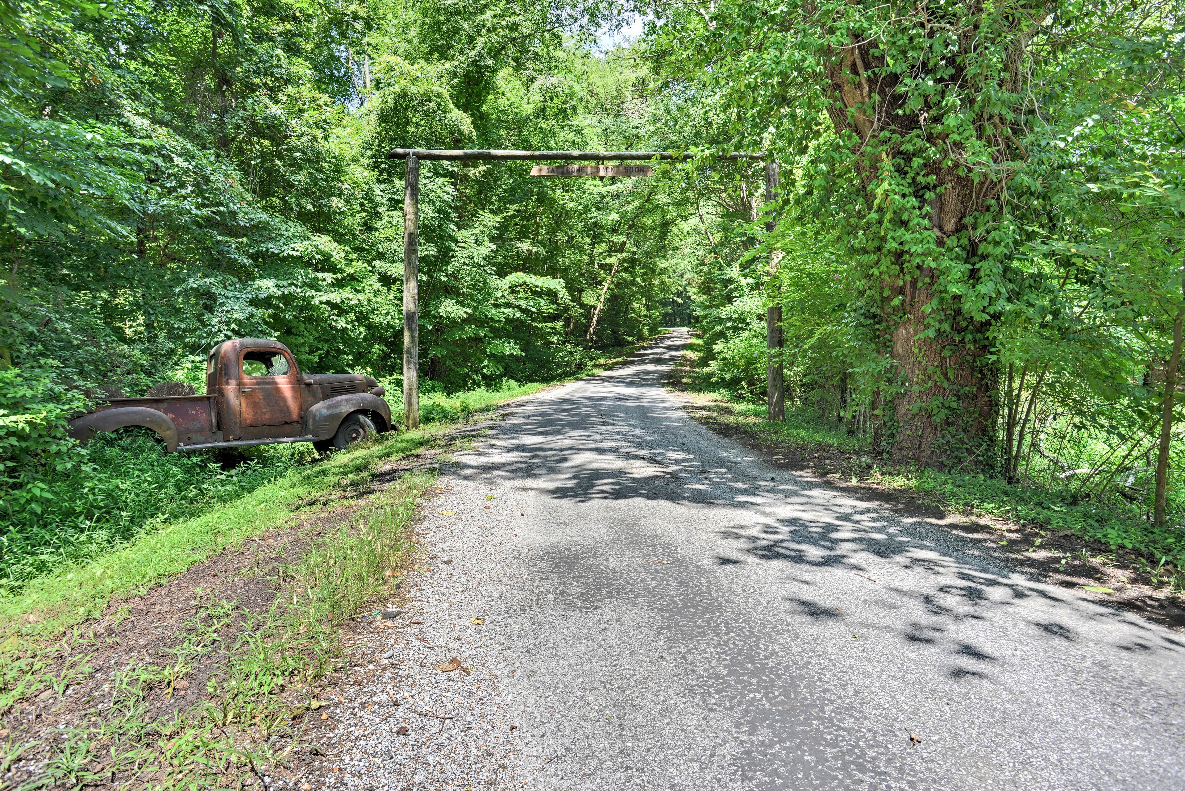 A rural road flanked by dense greenery and an old truck parked on the side.