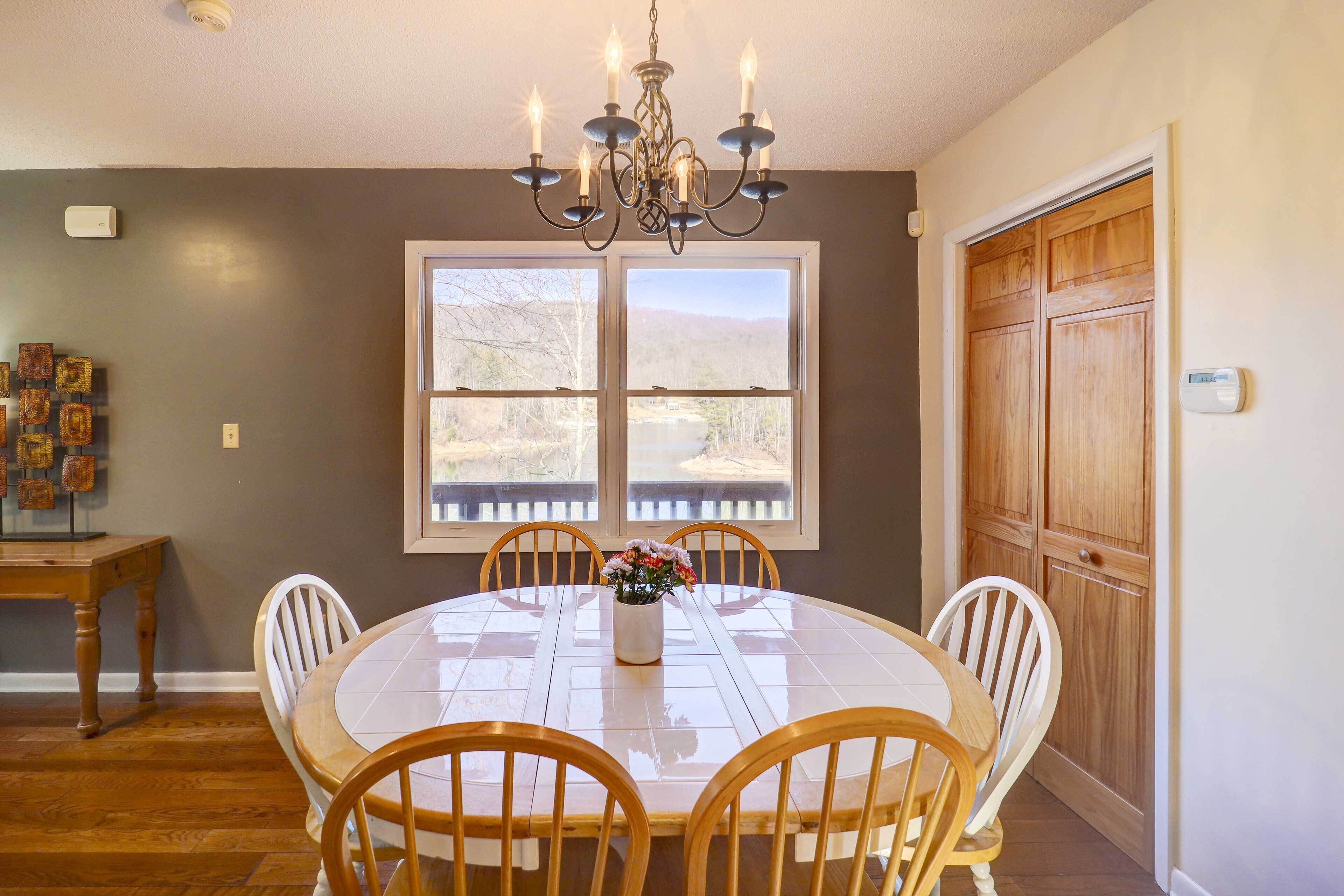A dining area with a round table, six chairs, a chandelier, a window with a view, and a wooden door.