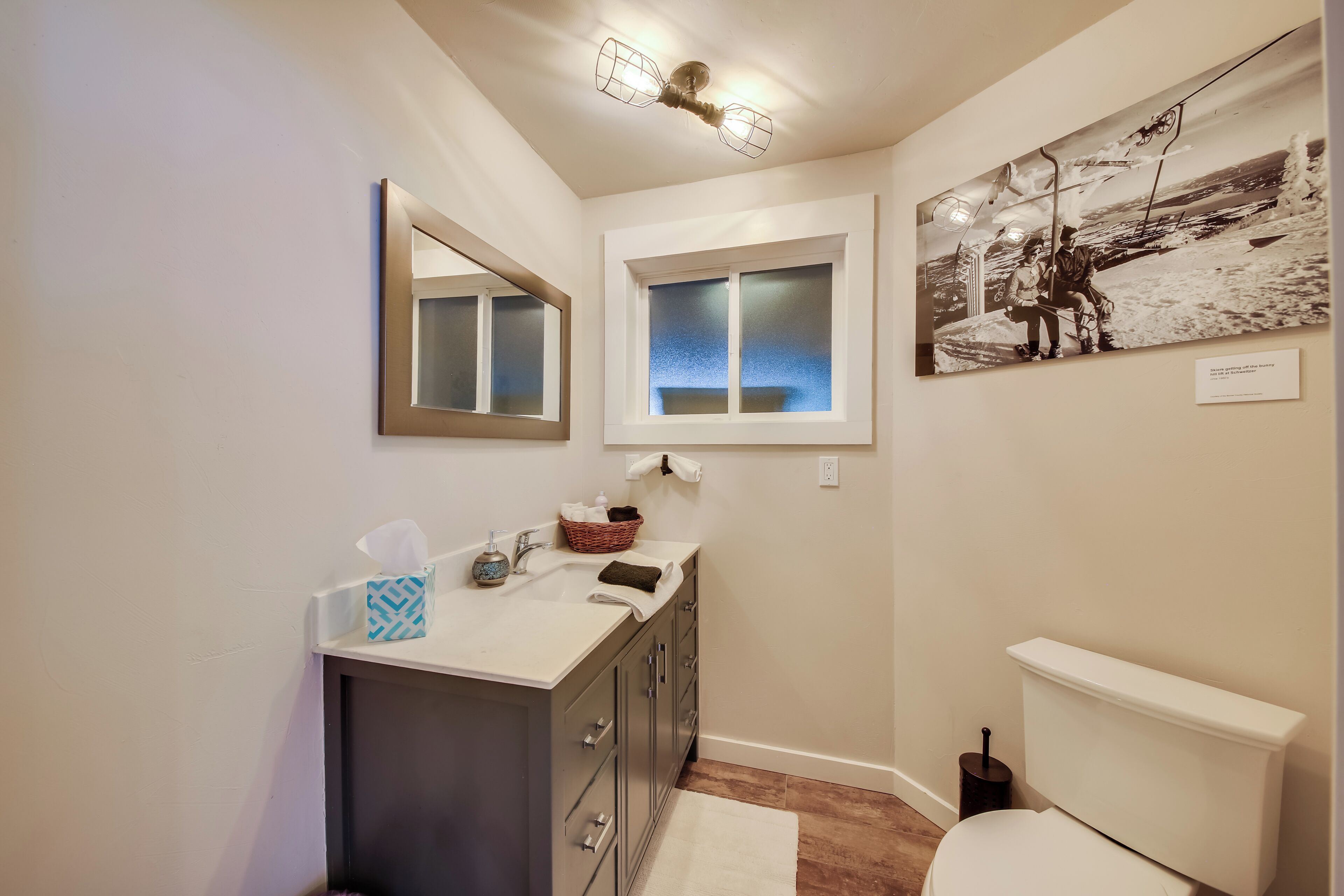 A bathroom with a large mirror, a window, a sink with toiletries, and a toilet.