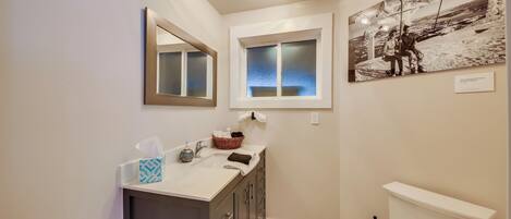 A bathroom with a large mirror, a window, a sink with toiletries, and a toilet.