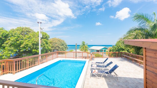 Ein Swimmingpool mit Holzterrasse und Liegestühlen, umgeben von einem Holzzaun und Palmen, mit Blick auf den Ozean.