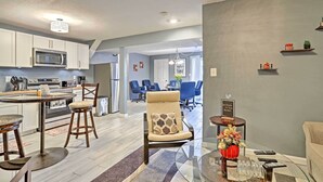 A modern kitchen with white cabinets, a stainless steel microwave, and a dining area with a round table and bar stools. There is a living room with a sofa and a coffee table with a vase and remote controls.