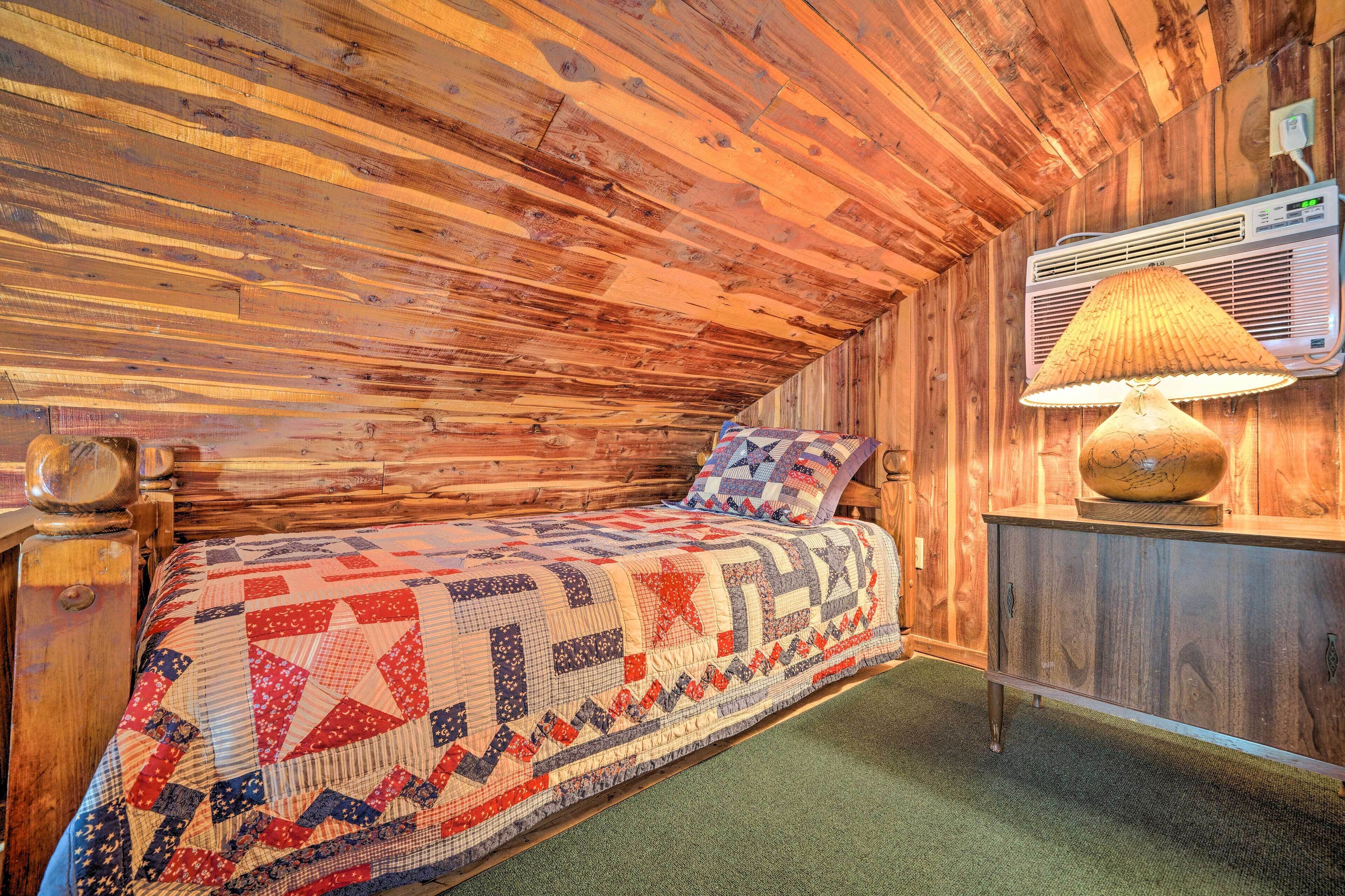 A cozy room with a wooden ceiling, a quilted bed, a wooden nightstand, and a wall-mounted air conditioner.