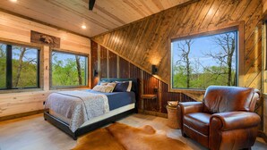 Une chambre à coucher avec un lit, un fauteuil en cuir, un tapis en peau de vache et de grandes fenêtres donnant sur la forêt.