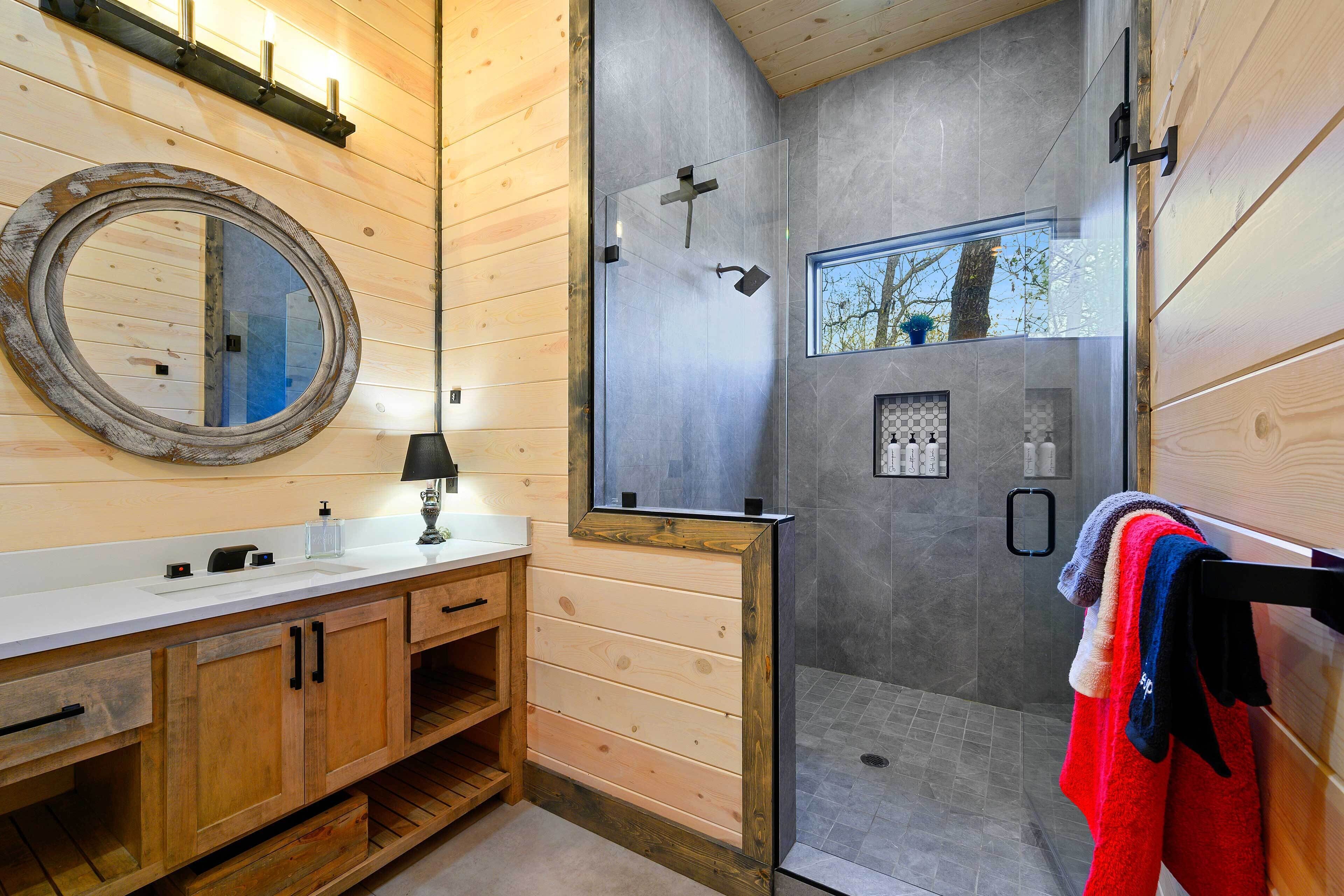 A modern bathroom with wooden paneling, a round mirror, a glass shower enclosure, and a window providing natural light.