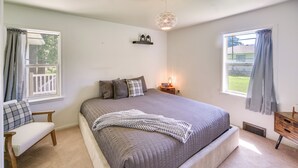 Un dormitorio con una cama grande, dos mesitas de noche, un pequeño tocador, una ventana con cortinas, una lámpara colgante y un estante sobre la cama.