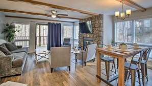 A spacious living room with a stone fireplace, wooden beams, a ceiling fan, and a dining area with metal chairs and a wooden table. - Mount Ida Condo - Walk to Lake Ouachita! (Mount Ida)