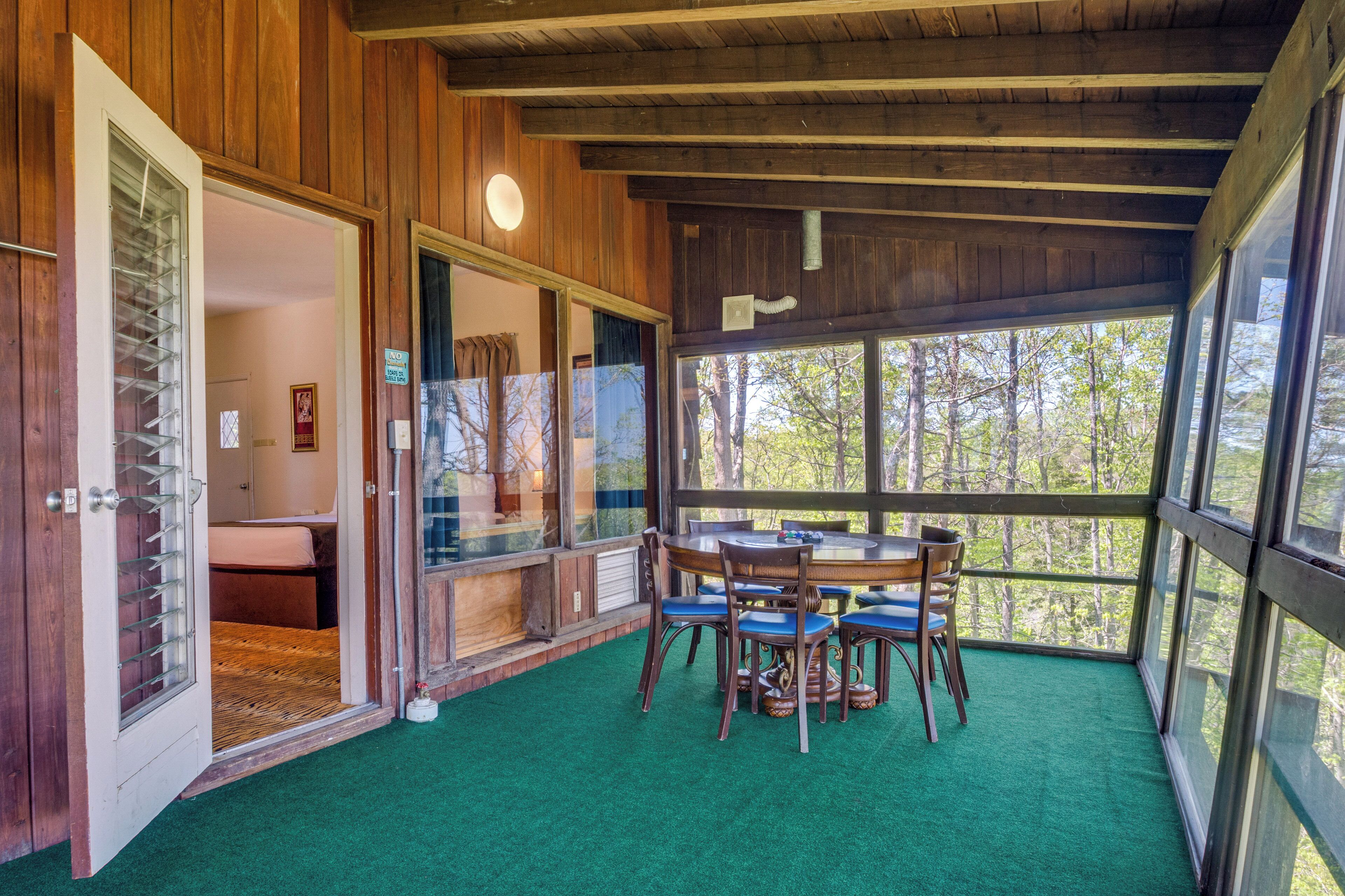 A room with a wooden ceiling and walls, a green carpet, a round table with chairs, and large windows offering a view of trees.