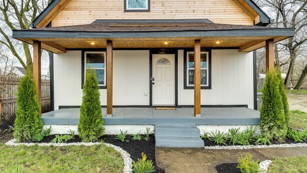 Une maison à deux étages, avec une façade en bois, un porche à l’avant et un petit jardin agrémenté d’arbustes et de plantes.