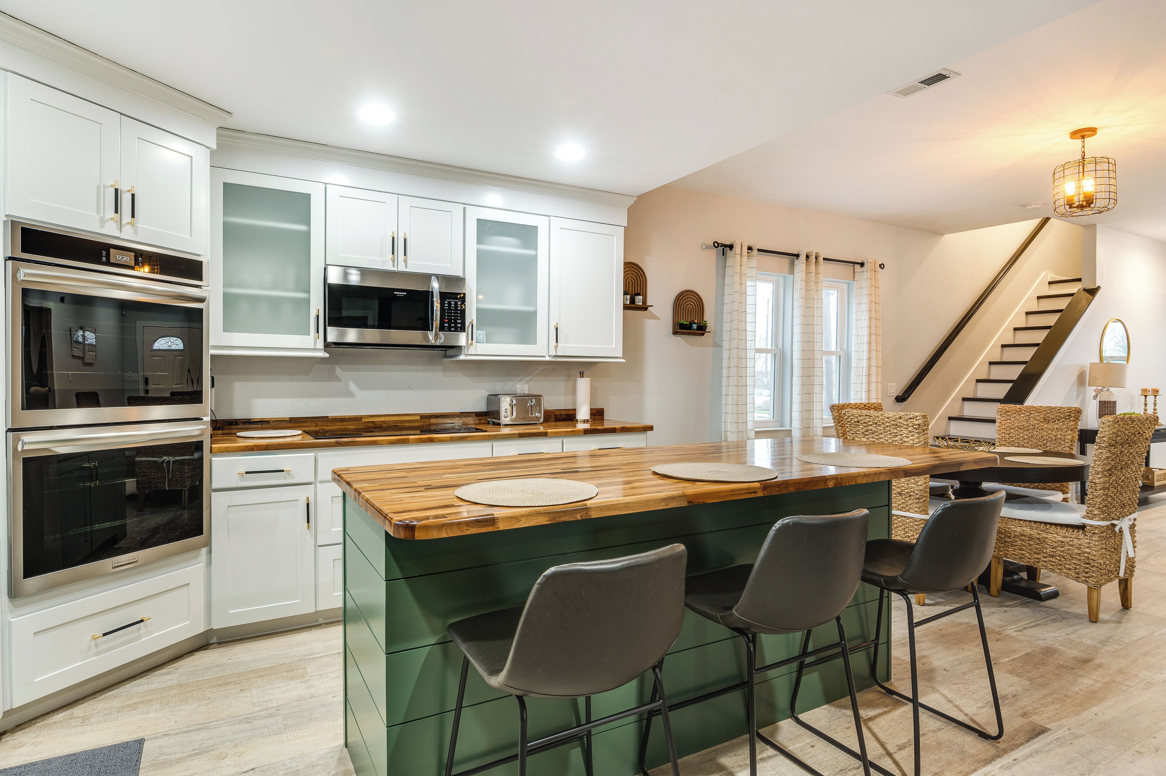 Una cocina moderna con armarios blancos, electrodomésticos de acero inoxidable, una isla de madera con taburetes altos y una escalera que sube a otro nivel.