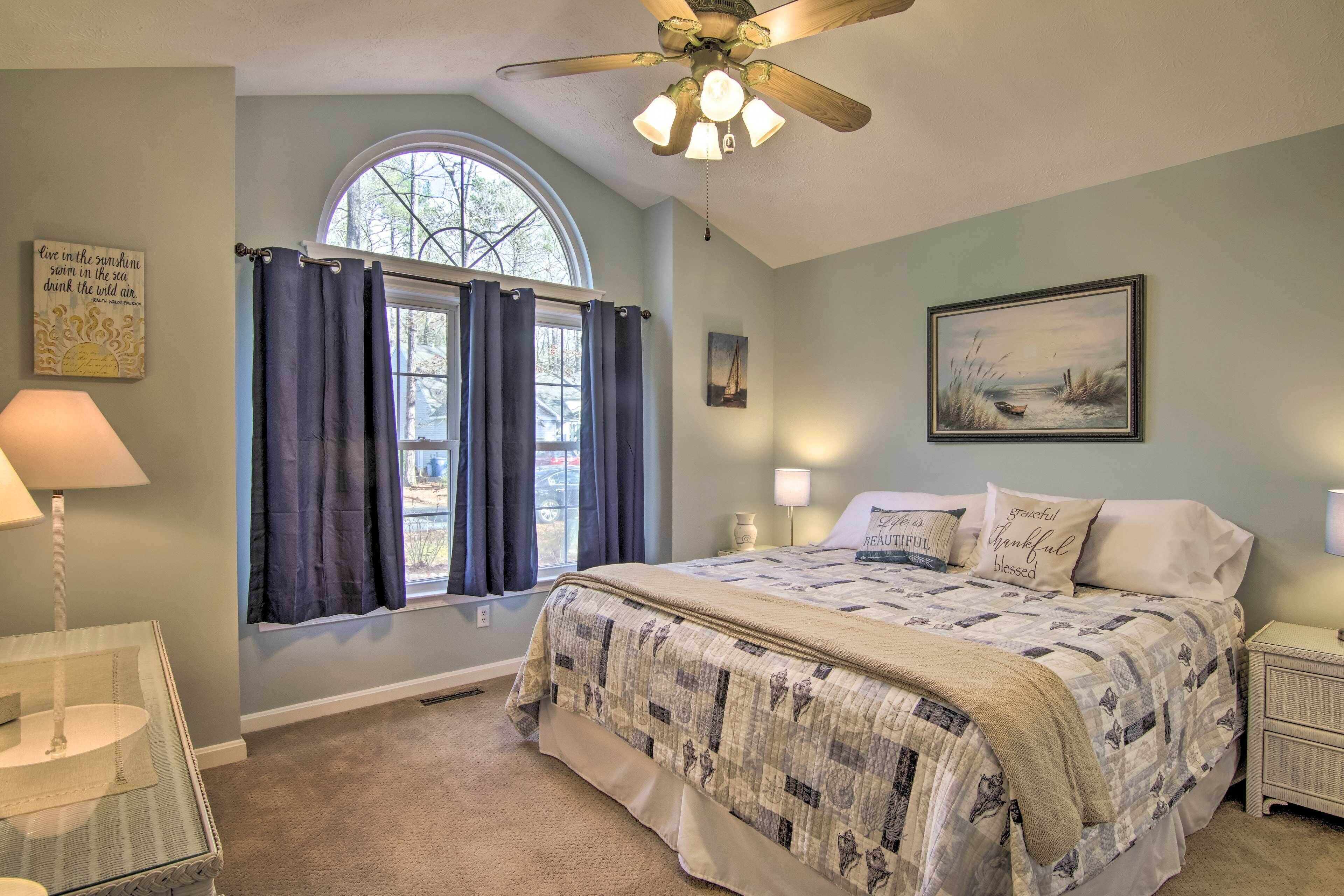 Um quarto com uma cama grande, um ventilador de teto, uma janela com cortinas, uma mesa de cabeceira com abajur e um quadro na parede.