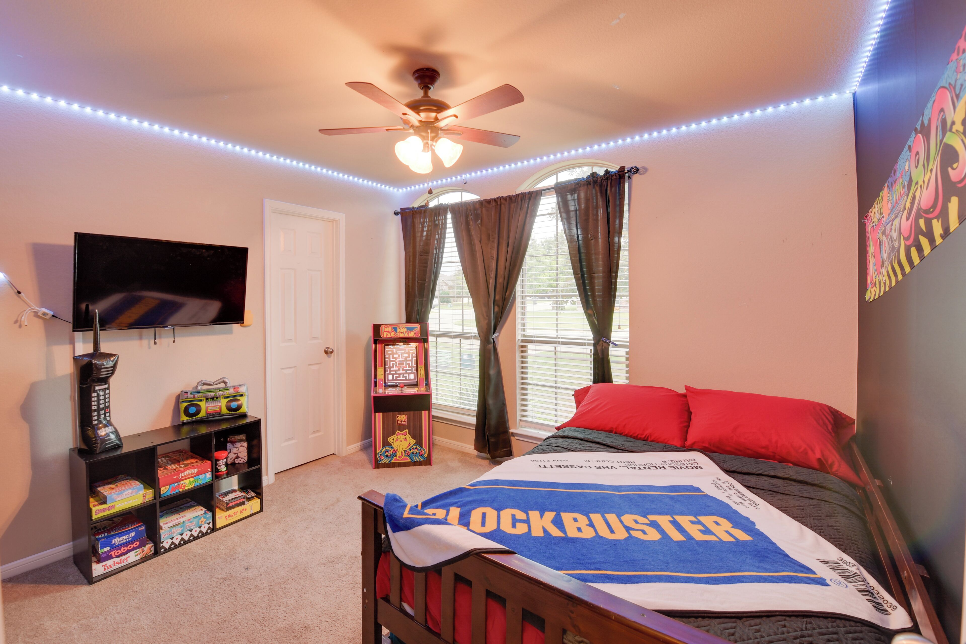 A bedroom with a bed, a ceiling fan, a TV, a bookshelf, and a window with curtains.