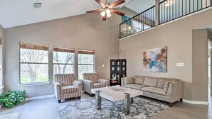 A spacious living room with a high ceiling, a ceiling fan, a beige sofa, two patterned armchairs, a coffee table, and a large window with blinds. - Peaceful Austin Home w/ Private Backyard! (Austin)