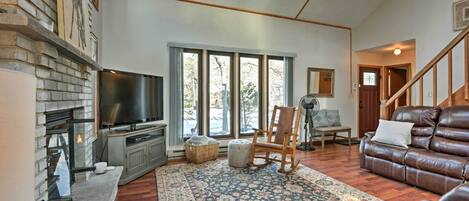 Un salon avec une cheminée, un téléviseur, un canapé en cuir, un fauteuil à bascule et une grande fenêtre donnant sur les arbres.