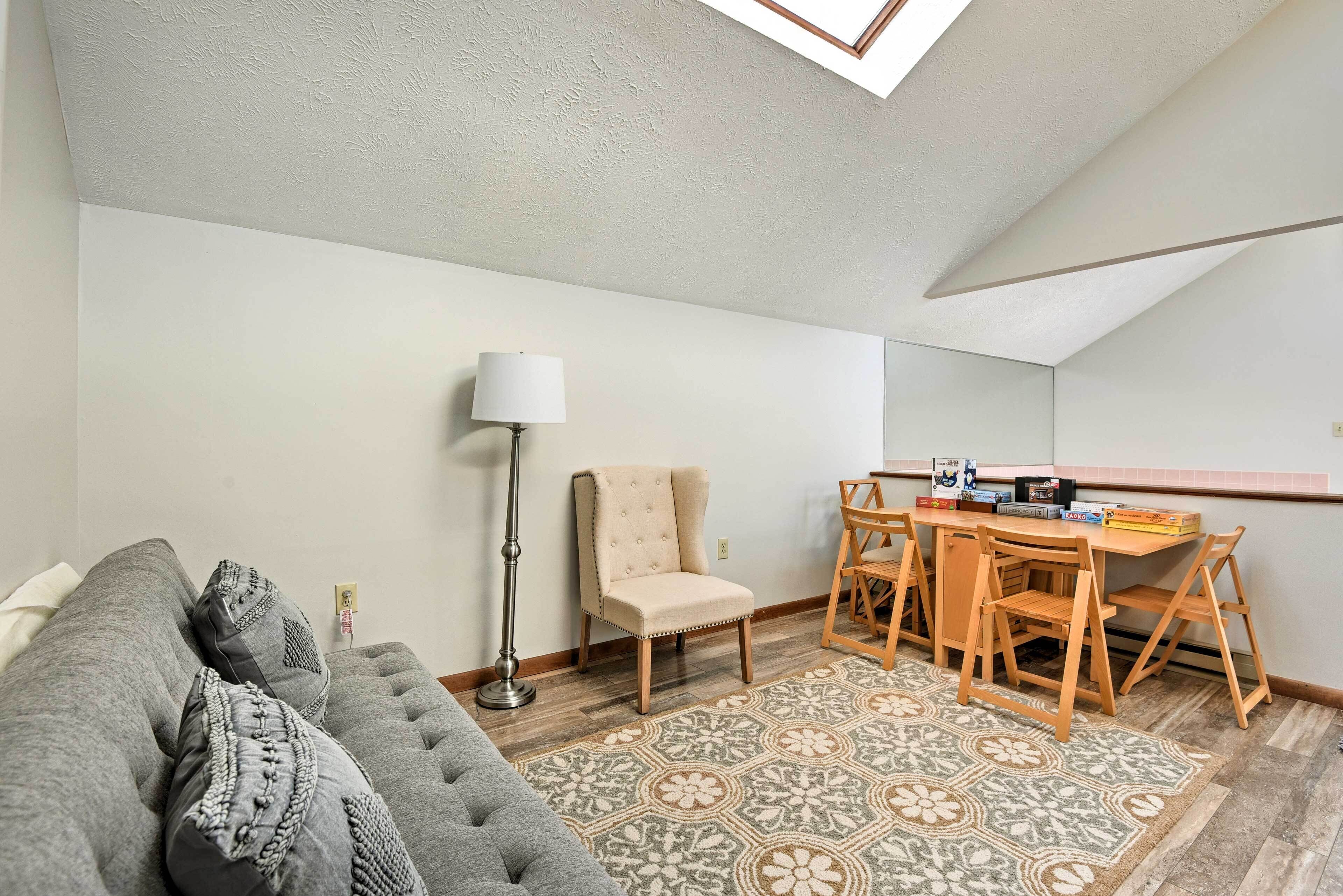A cozy living room with a grey sofa, a beige armchair, a wooden dining table with chairs, a patterned rug, and a floor lamp.