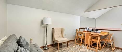 A cozy living room with a grey sofa, a beige armchair, a wooden dining table with chairs, a patterned rug, and a floor lamp.