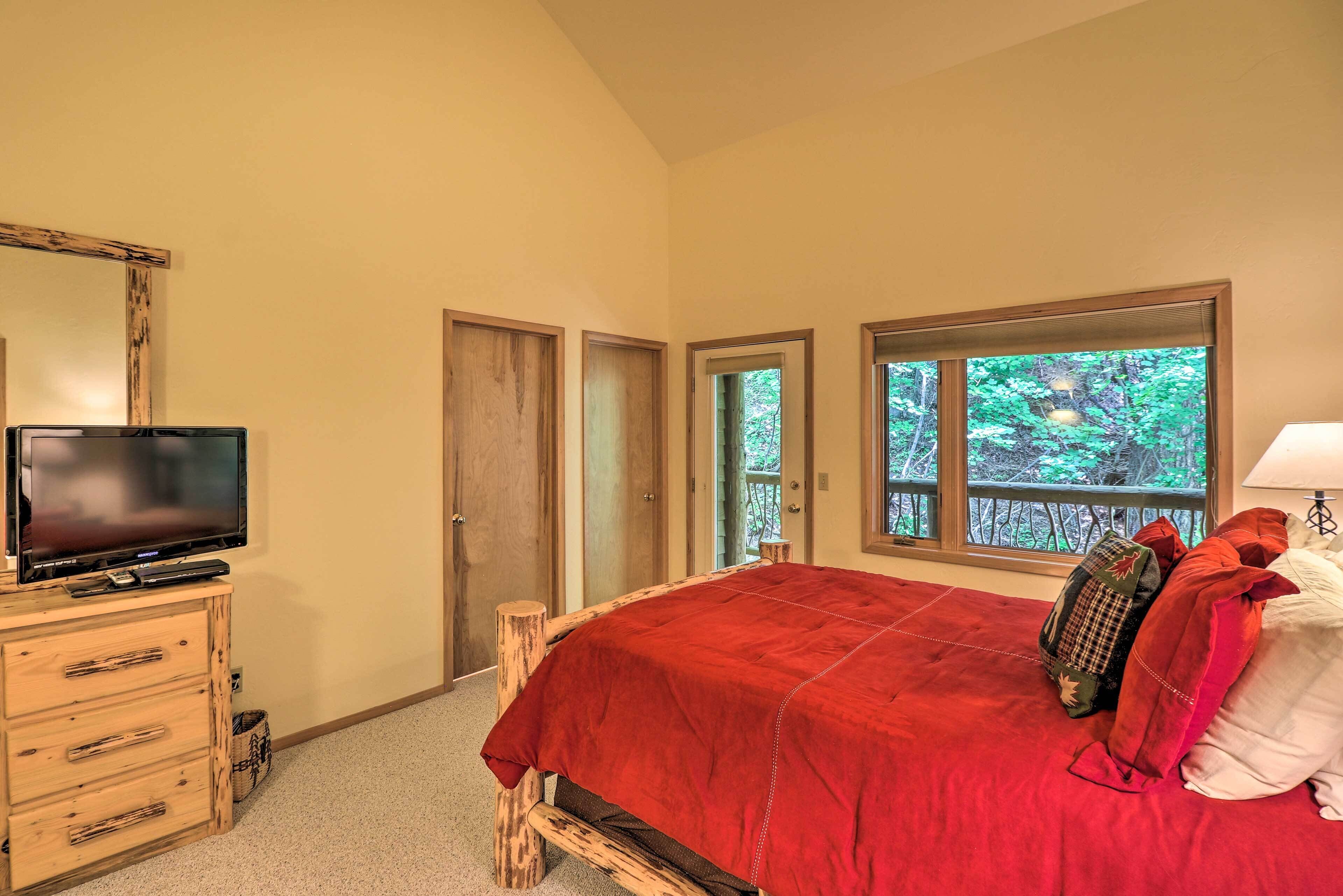 A bedroom with a bed, a TV on a wooden dresser, and a window with a view of trees.