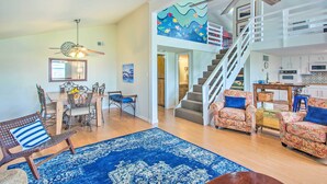 A spacious living area with a dining set, patterned sofas, a blue rug, a staircase, and a kitchen area.