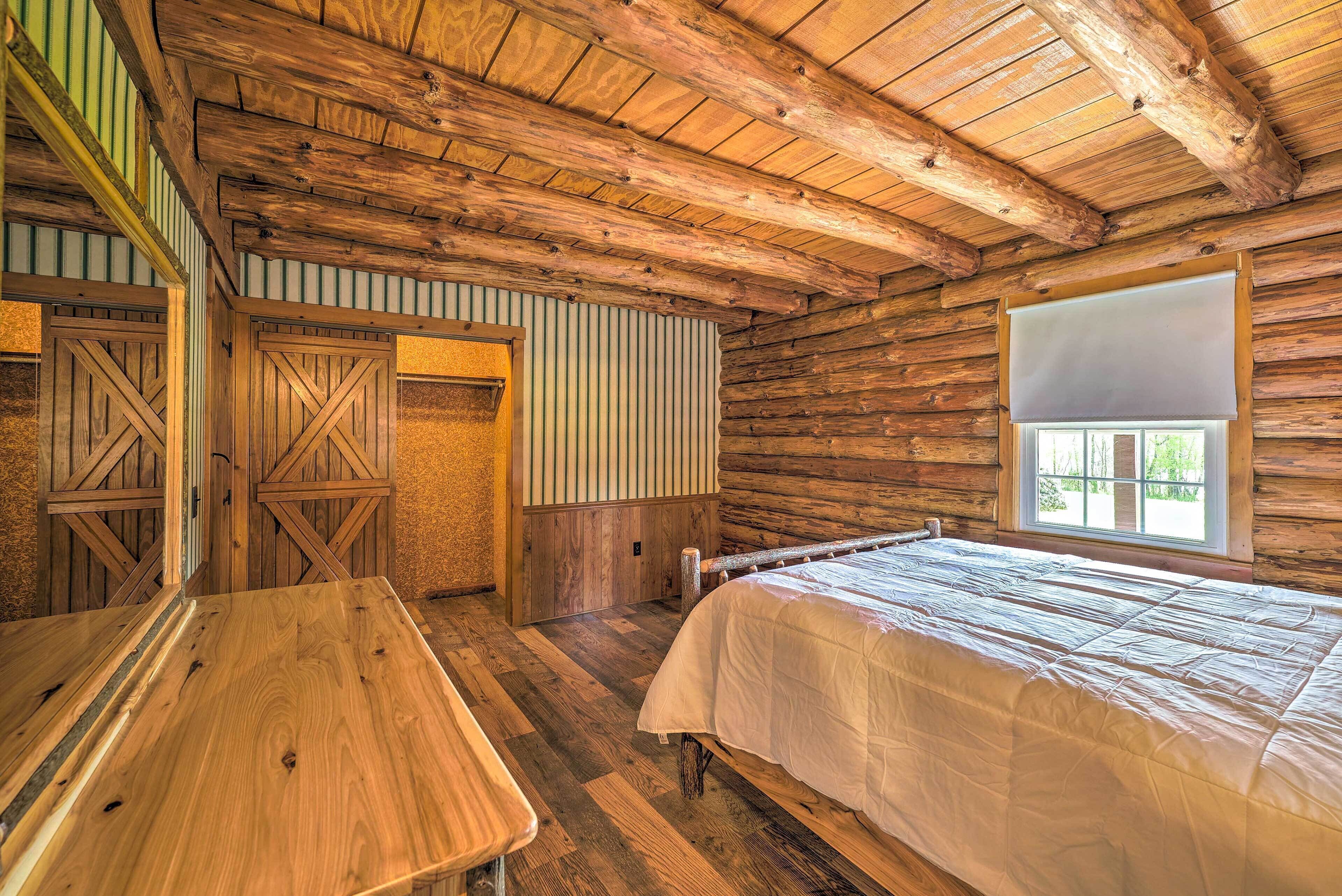 Ein Blockhauszimmer mit Holzboden, einem Bett mit weißer Bettdecke, einem Holztisch und einem Fenster mit Blick auf Grünfläche.