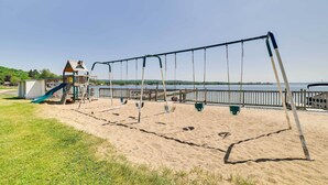 A playground with swings and a slide, a small building, and a view of the water. - Pool + Hot Tub Access! Portage Lake Condo w/ Deck (Onekama)