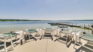 A deck with sun loungers overlooking a calm lake, a dock, and distant hills. - Pool + Hot Tub Access! Portage Lake Condo w/ Deck (Onekama)