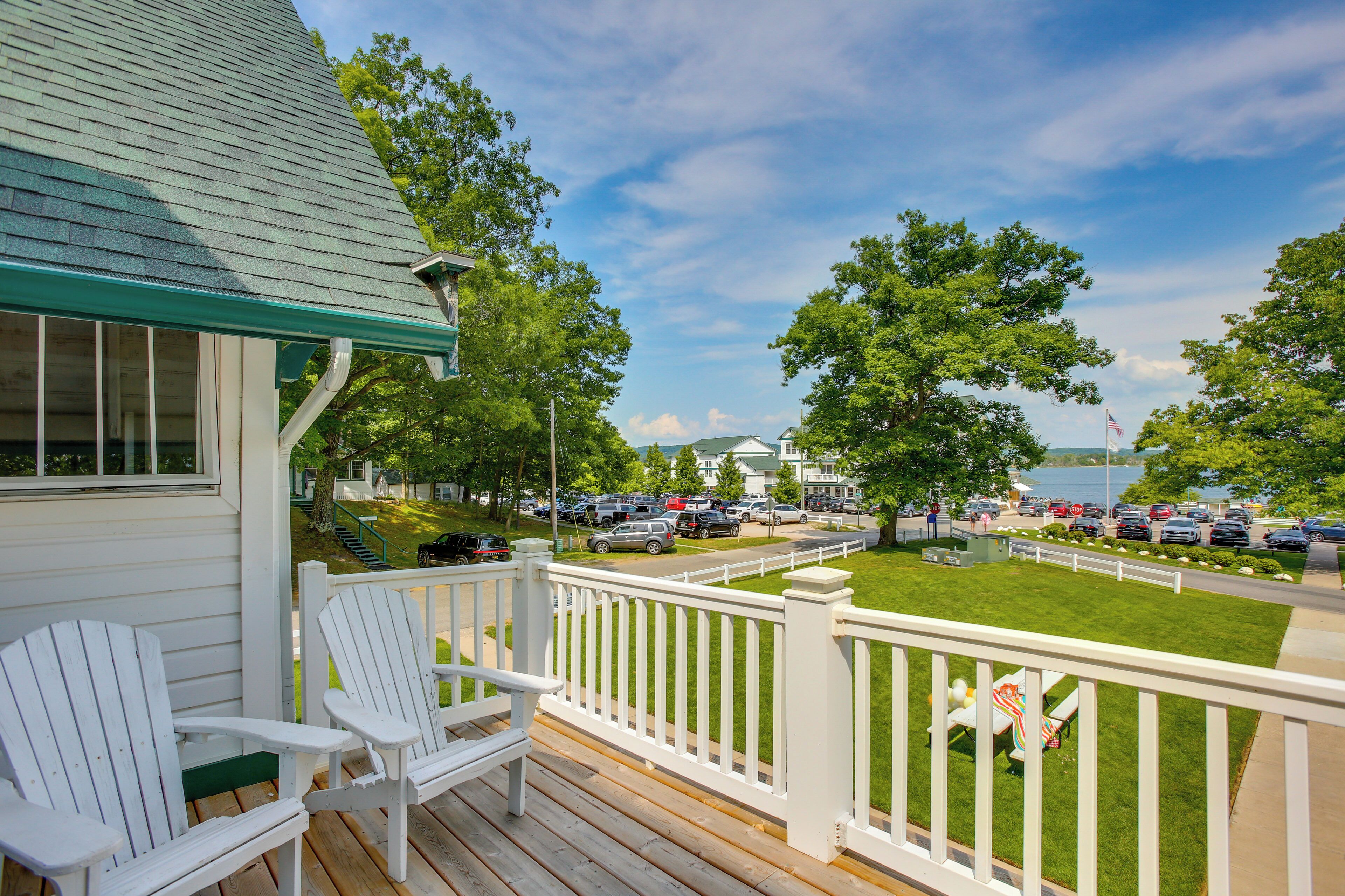 A wooden deck with white chairs, a view of a parking lot, and a lake in the background.