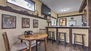 Un espace repas avec une table et des chaises en bois, un bar avec des tabourets, des tableaux encadrés accrochés au mur et un téléviseur fixé au-dessus du bar.