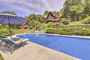 A swimming pool with lounge chairs and umbrellas, surrounded by a wooded area and a large house with a balcony. - Private Guest House w/ Deck + Spectacular Views! (Montville)