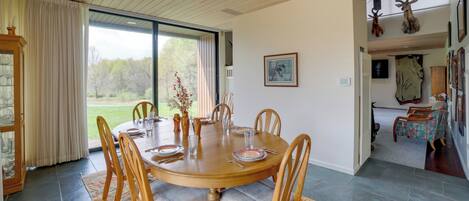 A dining area with a round wooden table, six chairs, and a glass-top table. There is a large window with curtains, a view of greenery outside, and a door leading to another room with a patterned rug and a deer head on the wall.