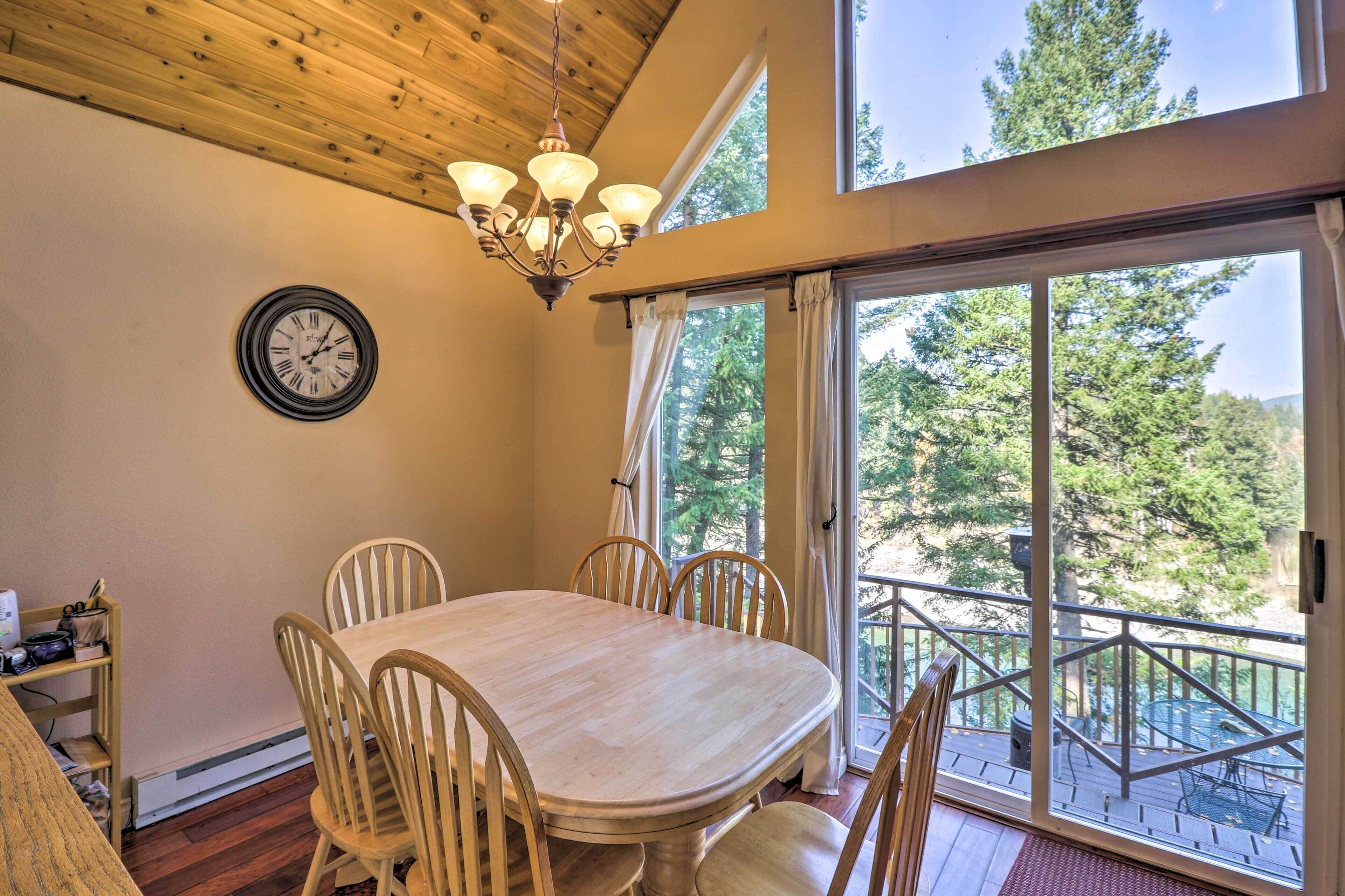 A dining area with a wooden table and chairs, a chandelier, a clock, and a large window with a view of trees.